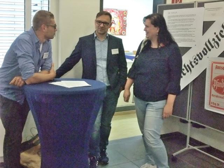 Gerichtsvollzieher im Gespräch Gerichtsvollzieher im Gespräch, 3 Personen an einem Stehtisch vor einer Info-Tafel